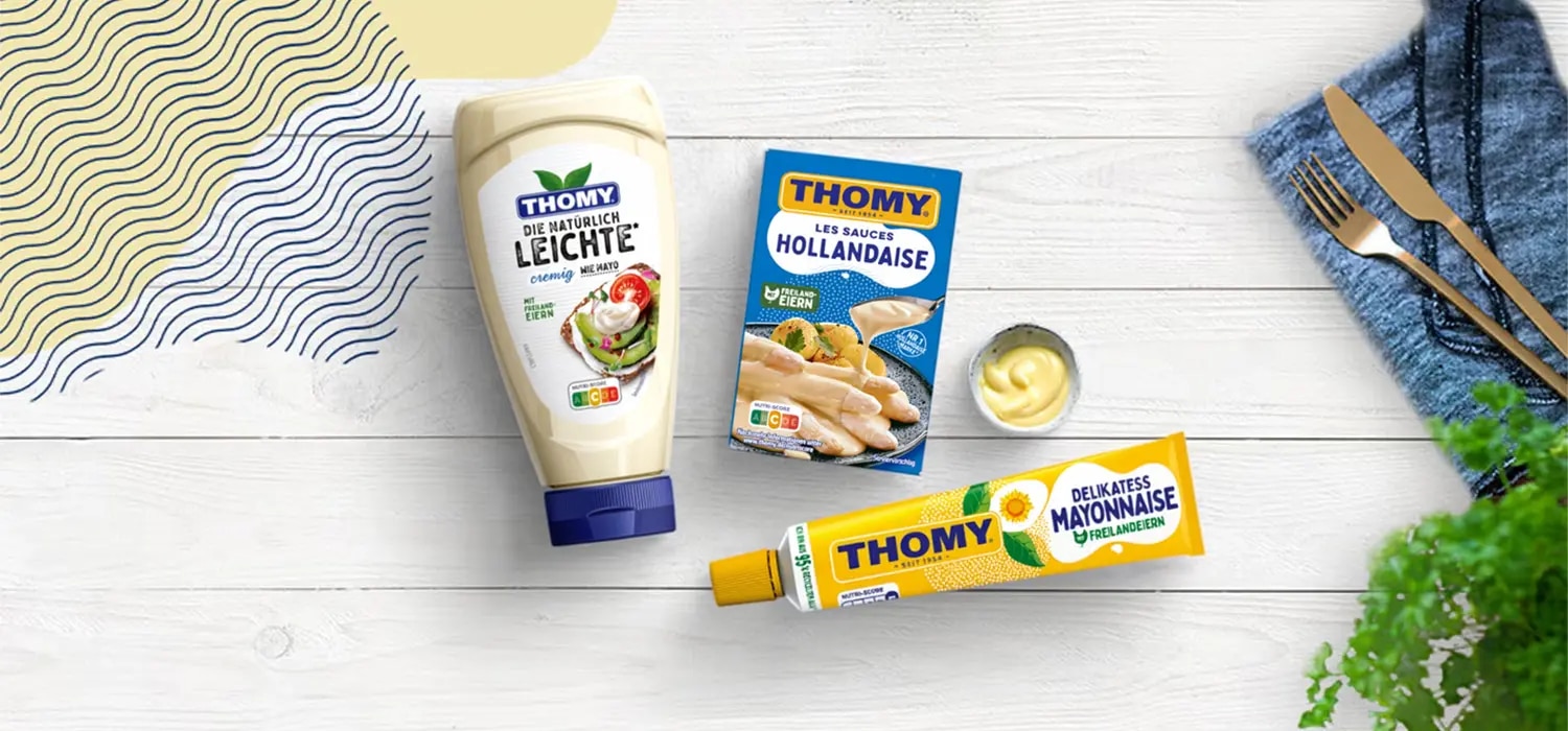 Thomy Produkte: Mayonnaise Flasche, Hollandaise Packung und Tube auf hellem Holztisch mit Besteck und Dip